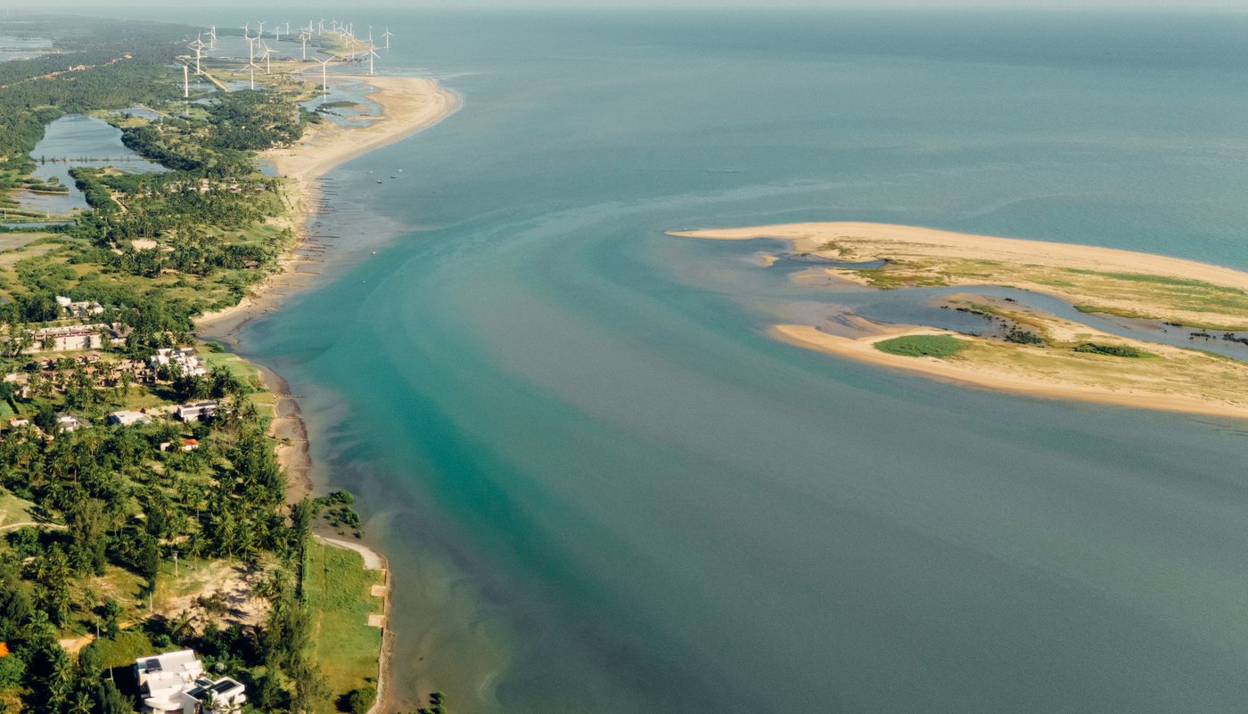 Vista aérea da costa com dunas, rio e parque eólico ao fundo